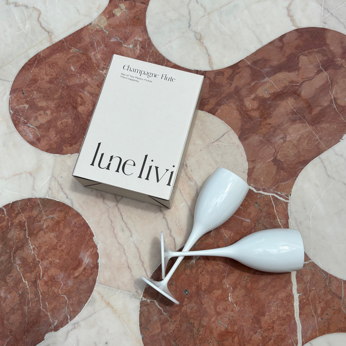 A boxed set of two white champagne flutes on a patterned brown and cream marble floor. The box is labeled 'lune livi' and 'Champagne Flutes'.