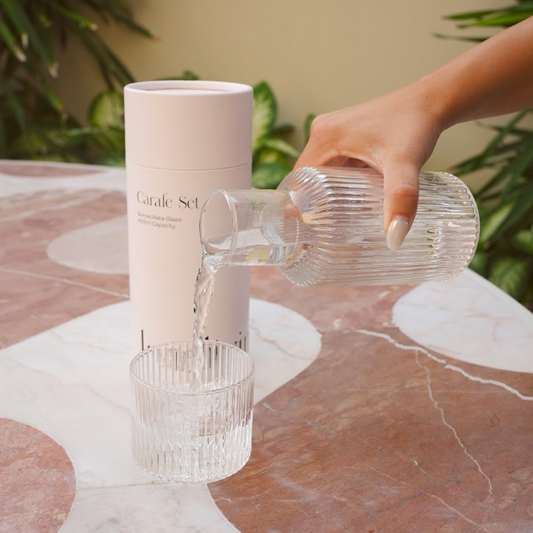 A hand pours water from a ribbed clear glass carafe into a matching ribbed glass tumbler, with a tall white cylindrical package standing behind them on a marble table and green plants in the background.
