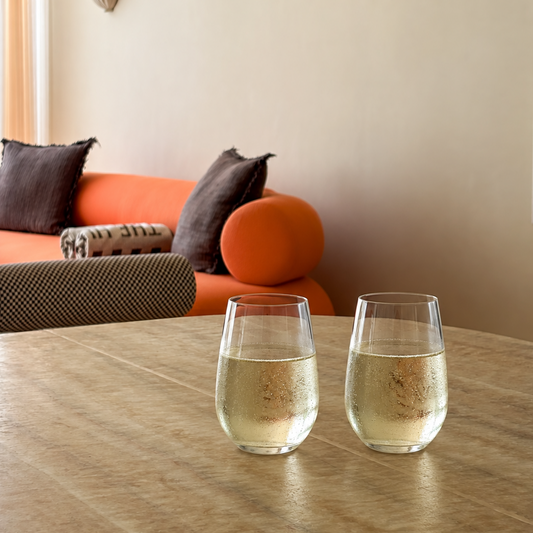 Two clear stemless wine glasses filled with a light golden beverage, placed on a wooden table in a warm, modern living room with an orange sofa in the background.