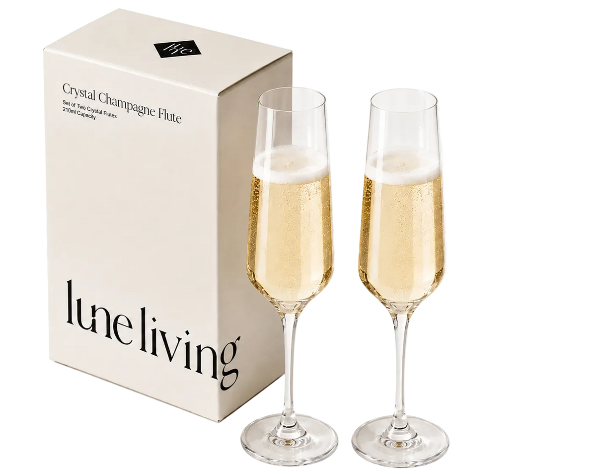 Two clear crystal champagne flutes filled with sparkling champagne, shown beside a cream-colored Luneliving product box on a white background.