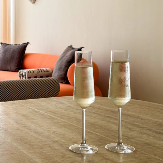 Two clear champagne flutes filled with pale golden bubbly drink, set on a round wooden table in a softly lit interior with an orange sofa and neutral wall in the background.